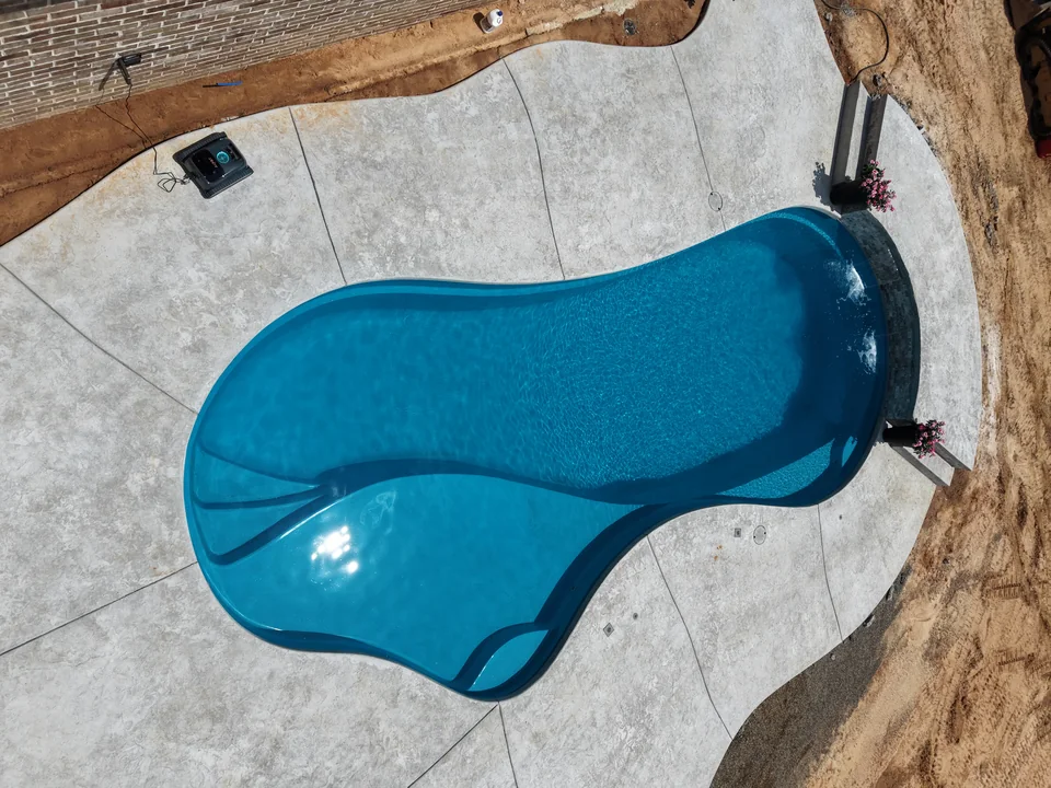 Freeform fiberglass pool with a large tanning ledge and a raised spillway wall, photographed before the yard was landscaped