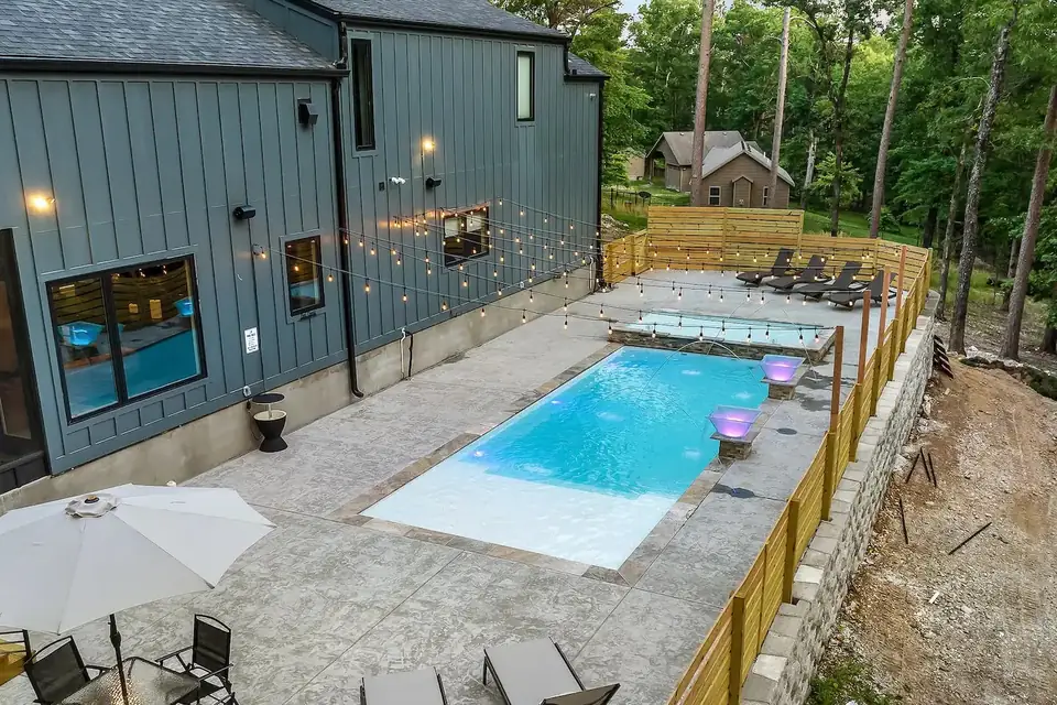 Fiberglass pool lit at dusk behind a dark board and batten home with string lights over the patio
