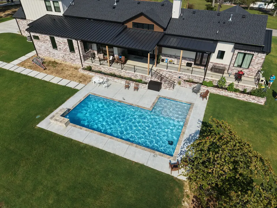 Rectangular fiberglass pool with a tanning ledge behind a modern farmhouse with a metal roof