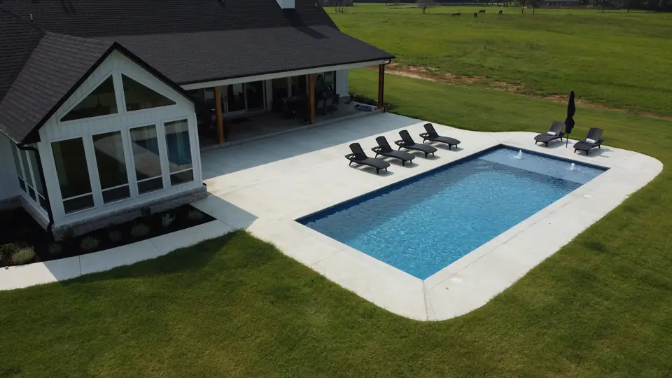 Rectangular fiberglass pool with deck jets and a wide concrete deck behind a white farmhouse