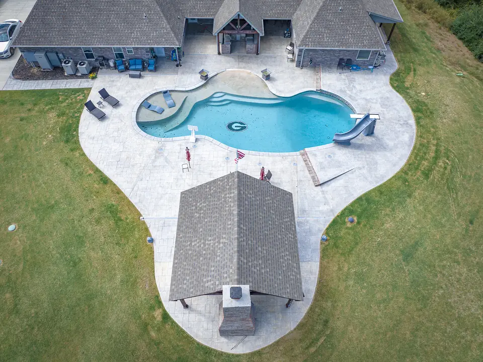 Large freeform fiberglass pool with a slide and a covered pavilion on a stamped concrete deck