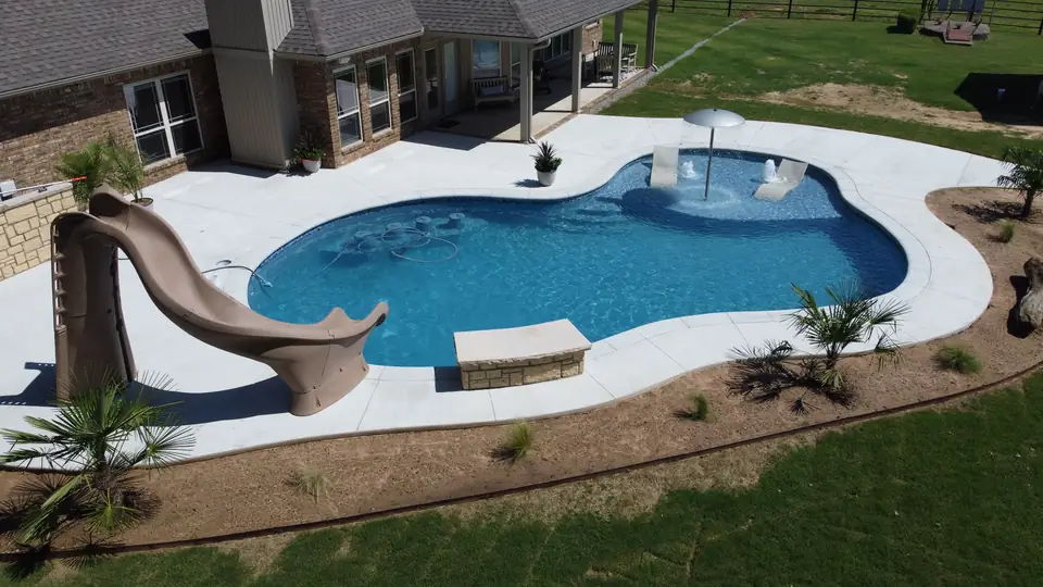 Freeform fiberglass pool with a slide, an umbrella water feature and a wide concrete deck